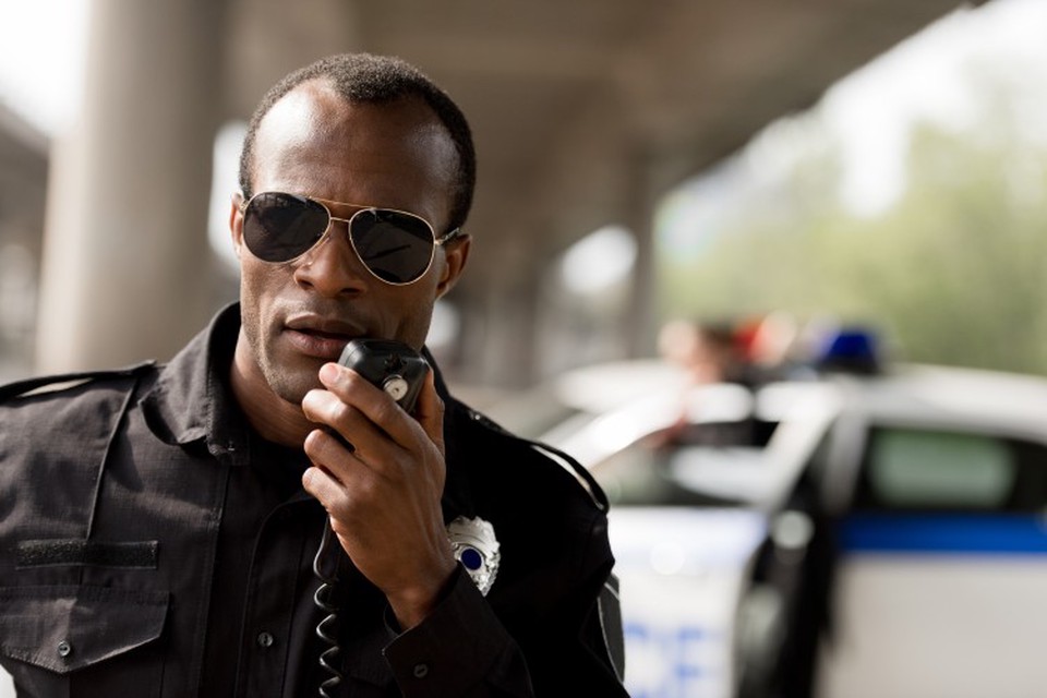 A law enforcement officer speaks on a radio with a police car in the background.