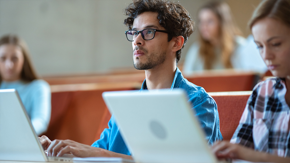 A student in a lecture hall looks up from working on his laptop.