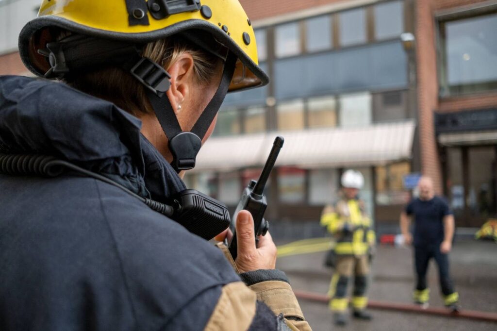 A homeland security and emergency preparedness professional wearing a helmet and bunker coat speaks into a walkie-talkie.