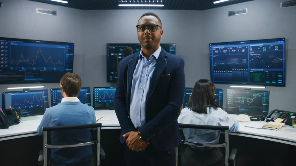 A man in a suit stands in front of a data analysis control room with multiple screens.