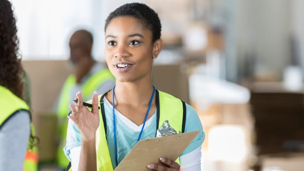 A woman in a safety vest holds a clipboard and gestures while speaking in a workplace setting.