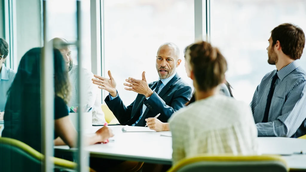 A man in a suit gestures as he leads a meeting with people sitting around a table.
