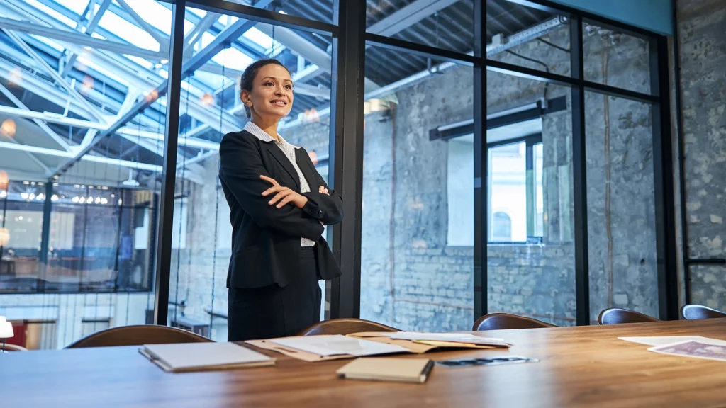 A woman in a black suit stands in a modern meeting room with large glass windows and a wooden table.