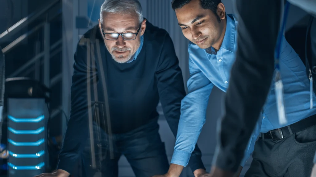 Two men lean over a lit table in a high-tech office setting.