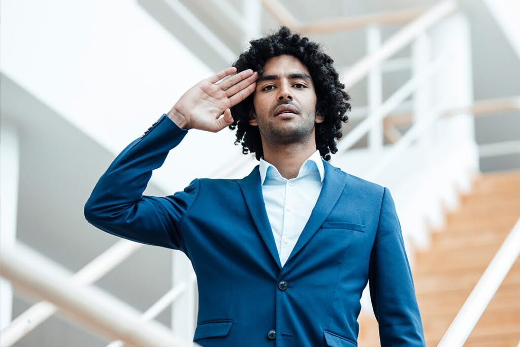 A man in a blue suit standing indoors, looking forward and saluting.