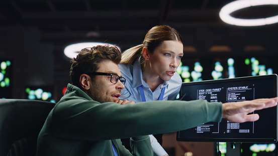 Two professionals analyze code on a computer screen in a modern, high-tech office; one points at the monitor while the other observes closely.
