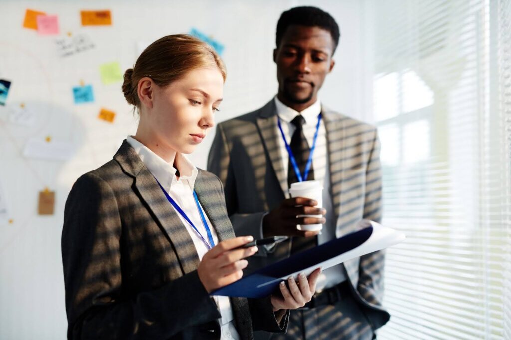 Two professionals in business attire review a document together in a bright office, with notes and photos pinned on the wall in the background.