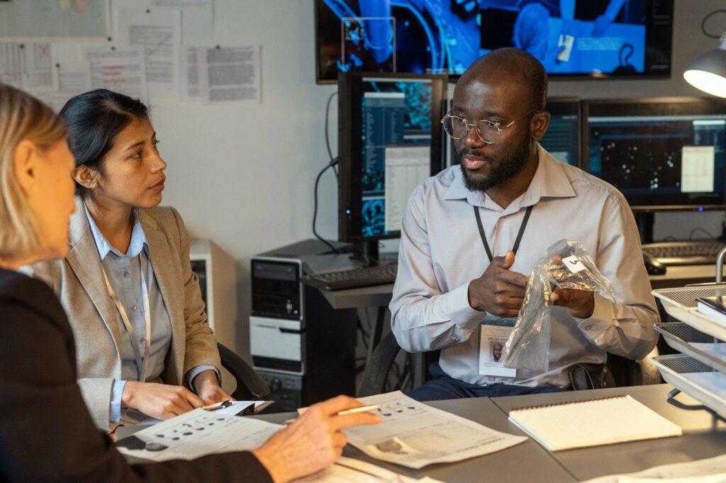 Three professionals seated at a desk engage in discussion while examining evidence in a plastic bag, with computer monitors and documents in the background.