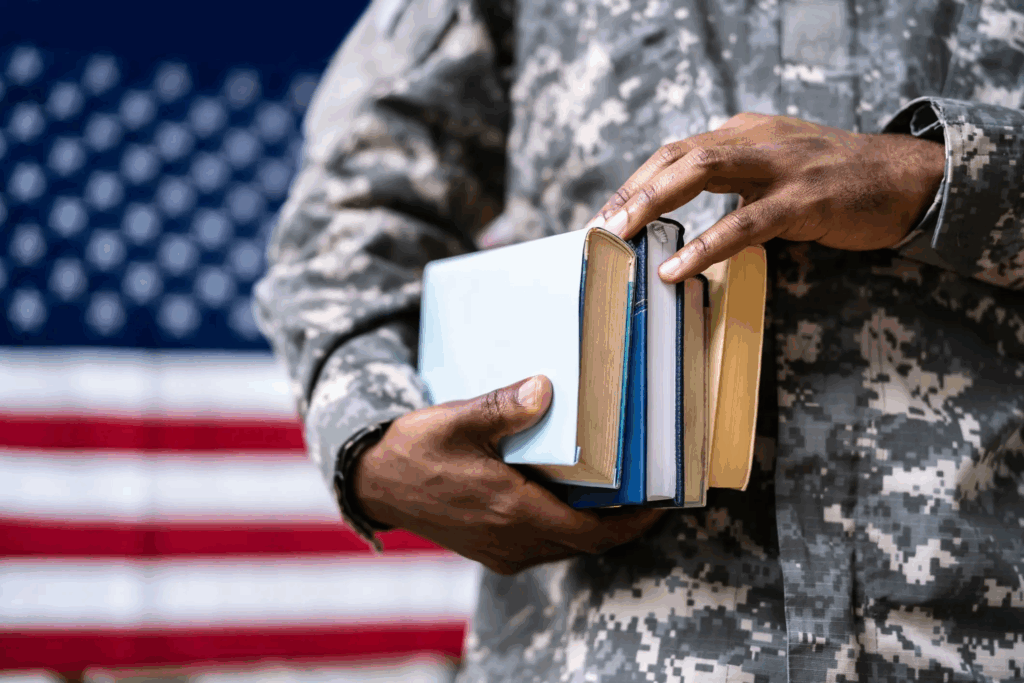 A closeup of a military person's hands holding a stack of books as he stands in front of an American flag.