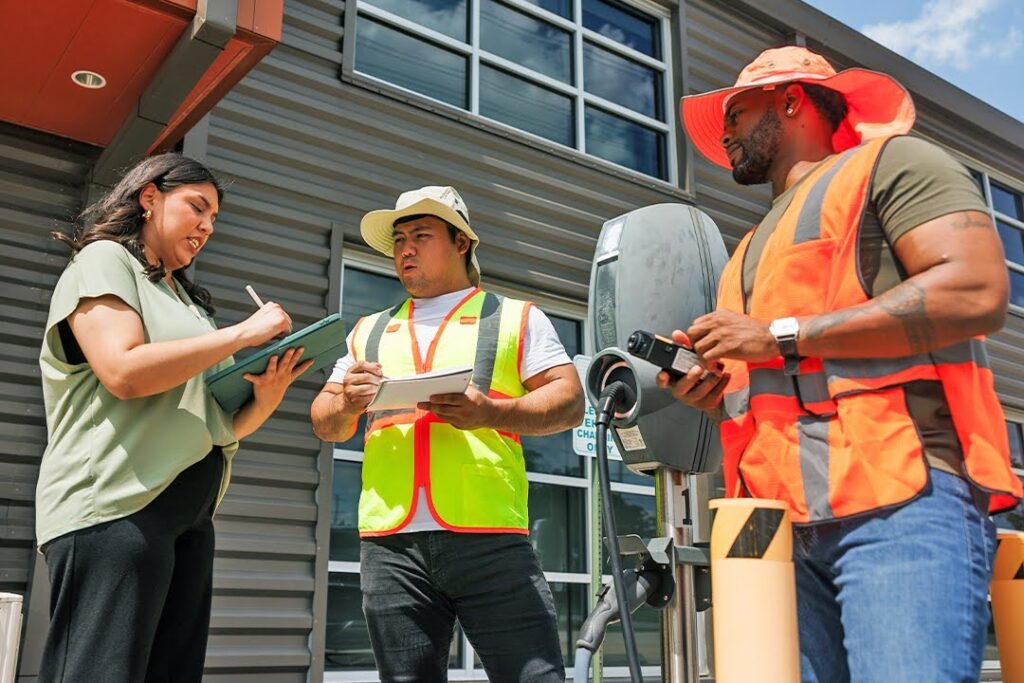 Three engineers in safety vests inspect an electric vehicle charging station.